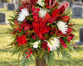Könnte beinhalten: Ein Blumenarrangement in einer bronzefarbenen Vase mit roten Weihnachtssternen, weißen Chrysanthemen, grünem Blattwerk, roten Beeren und rot-weiß gestreiften Zuckerstangen-Akzenten, platziert auf einem Granitsockel.