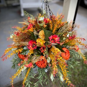 K&ouml;nnte beinhalten: Ein h&auml;ngendes Blumenarrangement mit einer dichten Ansammlung k&uuml;nstlicher Blumen und Bl&auml;tter in Orange-, Gelb-, Rot- und Gr&uuml;nt&ouml;nen. Der Strau&szlig; enth&auml;lt runde orangefarbene Pfingstrosen, rote G&auml;nsebl&uuml;mchen und gelbe Chrysanthemen.