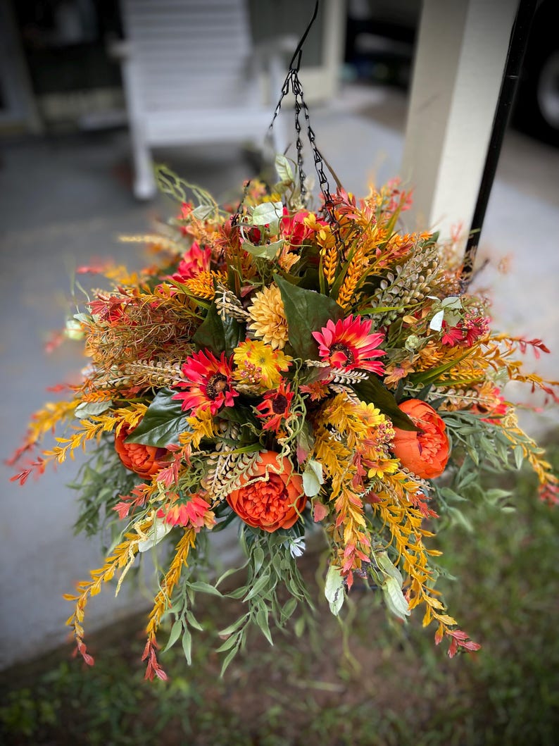 K&ouml;nnte beinhalten: Ein h&auml;ngendes Blumenarrangement mit einer dichten Ansammlung k&uuml;nstlicher Blumen und Bl&auml;tter in Orange-, Gelb-, Rot- und Gr&uuml;nt&ouml;nen. Der Strau&szlig; enth&auml;lt runde orangefarbene Pfingstrosen, rote G&auml;nsebl&uuml;mchen und gelbe Chrysanthemen.