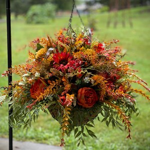 K&ouml;nnte beinhalten: Ein H&auml;ngekorb gef&uuml;llt mit k&uuml;nstlichem Herbstlaub, darunter dunkelrote Sonnenblumen, orangefarbene Pfingstrosen, gelbe Chrysanthemen sowie verschiedene gr&uuml;ne und rostfarbene Blattzweige, aufgeh&auml;ngt an einer d&uuml;nnen Metallkette an einem schwarzen Haken.