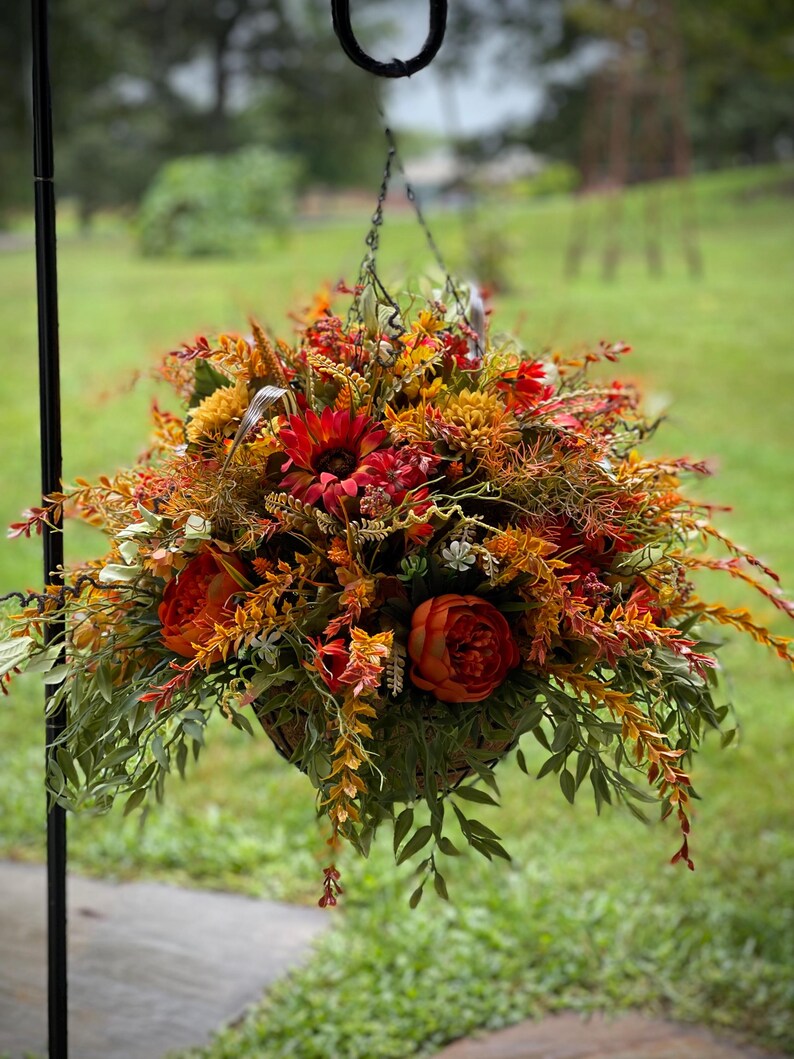 K&ouml;nnte beinhalten: Ein H&auml;ngekorb gef&uuml;llt mit k&uuml;nstlichem Herbstlaub, darunter dunkelrote Sonnenblumen, orangefarbene Pfingstrosen, gelbe Chrysanthemen sowie verschiedene gr&uuml;ne und rostfarbene Blattzweige, aufgeh&auml;ngt an einer d&uuml;nnen Metallkette an einem schwarzen Haken.