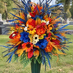 May include: A floral arrangement featuring vibrant blue, orange, and yellow artificial flowers, including dahlias and sunflowers, mixed with green foliage and tall blue grass-like stems, all contained within a dark green plastic cemetery vase.