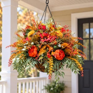 K&ouml;nnte beinhalten: Ein H&auml;ngekorb gef&uuml;llt mit k&uuml;nstlichem Herbstlaub, darunter leuchtend orangefarbene Pfingstrosen, gelbe Sonnenblumen, rote G&auml;nsebl&uuml;mchen und verschiedene gr&uuml;ne Zweige. Das Arrangement befindet sich in einem dunklen Drahtkorb an einer schwarzen Kette.