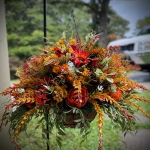 K&ouml;nnte beinhalten: Ein H&auml;ngekorb gef&uuml;llt mit k&uuml;nstlichen Herbstblumen und Blattwerk in Rot-, Orange- und Gelbt&ouml;nen. Das Arrangement besteht aus Sonnenblumen, Pfingstrosen und verschiedenen bl&auml;ttrigen Stielen, die in einem runden, braunen Korb stecken.