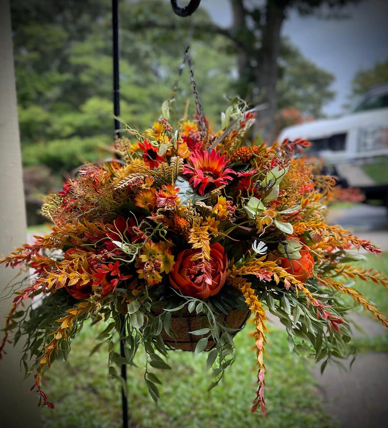 K&ouml;nnte beinhalten: Ein H&auml;ngekorb gef&uuml;llt mit k&uuml;nstlichen Herbstblumen und Blattwerk in Rot-, Orange- und Gelbt&ouml;nen. Das Arrangement besteht aus Sonnenblumen, Pfingstrosen und verschiedenen bl&auml;ttrigen Stielen, die in einem runden, braunen Korb stecken.