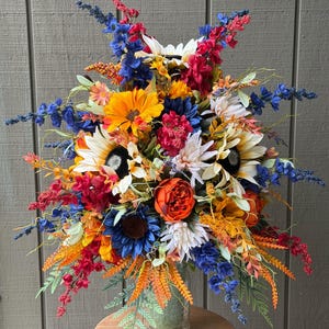 May include: A large, vibrant floral arrangement in a clear glass vase, featuring a mix of sunflowers, orange peonies, blue delphiniums, and red astilbe. The bouquet includes various green foliage and filler flowers, creating a dense, colorful display.