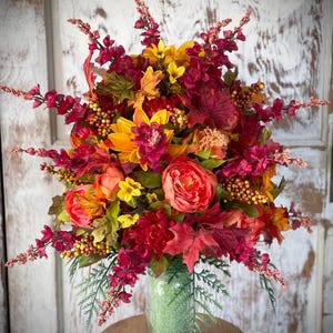 May include: A fall-themed floral arrangement in a clear glass jar. The arrangement features red, orange, and yellow flowers, along with green leaves and berries.