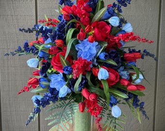 Könnte beinhalten: Ein Blumenarrangement aus künstlichen roten, dunkelblauen und hellblauen Blüten, darunter Rosen, Hortensien und hohe, spitze Blumen. Die Pflanzen stecken in einer klaren Glasvase mit grünem Steckschaum und bilden ein dichtes Arrangement.