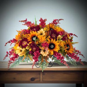 May include: A floral arrangement featuring bright yellow sunflowers, deep burgundy dahlias, and small red filler flowers mixed with green foliage. The bouquet is displayed in a black metal stand, creating a dense, rounded display of autumn colors.