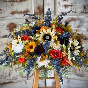 May include: A floral arrangement featuring sunflowers with white and yellow petals, deep red roses, and dark blue sprigs of lavender-like flowers. The bouquet includes green foliage and small yellow accents, creating a dense, rounded display of colors.