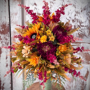 May include: A vibrant floral arrangement featuring sunflowers, dahlias, and various wildflowers in shades of yellow, orange, and burgundy. The bouquet is arranged in a clear glass jar wrapped in green fabric, placed on a wooden stool.
