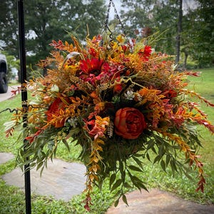 K&ouml;nnte beinhalten: Ein h&auml;ngendes Blumenarrangement mit einem dichten B&uuml;schel aus k&uuml;nstlichen Blumen und Bl&auml;ttern in den Farben Dunkelrot, gebranntes Orange, Goldgelb und Salbeigr&uuml;n. Der Strau&szlig; ist an einer d&uuml;nnen Metallkette befestigt.