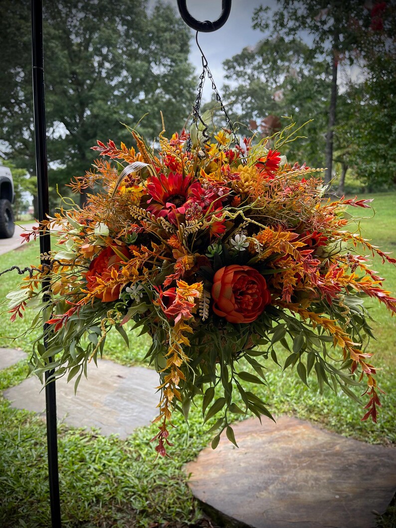 K&ouml;nnte beinhalten: Ein h&auml;ngendes Blumenarrangement mit einem dichten B&uuml;schel aus k&uuml;nstlichen Blumen und Bl&auml;ttern in den Farben Dunkelrot, gebranntes Orange, Goldgelb und Salbeigr&uuml;n. Der Strau&szlig; ist an einer d&uuml;nnen Metallkette befestigt.