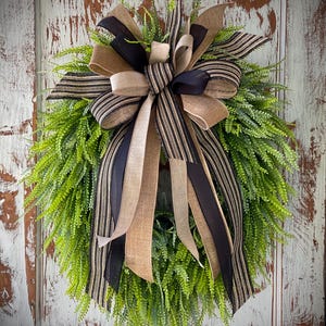 May include: A decorative wreath featuring a lush green base of faux foliage. The wreath is adorned with a large bow made of black, tan, and striped ribbons. The wreath is designed for door or wall decor.