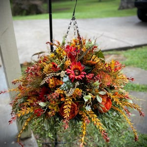 K&ouml;nnte beinhalten: Ein h&auml;ngendes Blumenarrangement mit einer dichten Ansammlung k&uuml;nstlicher Blumen und Bl&auml;tter in Rot-, Orange-, Gelb- und Gr&uuml;nt&ouml;nen. Der Strau&szlig; enth&auml;lt Sonnenblumen, Pfingstrosen und verschiedene bl&auml;ttrige Stiele, die an einer Metallkette h&auml;ngen.
