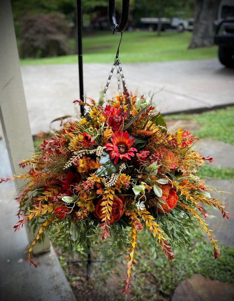 K&ouml;nnte beinhalten: Ein h&auml;ngendes Blumenarrangement mit einer dichten Ansammlung k&uuml;nstlicher Blumen und Bl&auml;tter in Rot-, Orange-, Gelb- und Gr&uuml;nt&ouml;nen. Der Strau&szlig; enth&auml;lt Sonnenblumen, Pfingstrosen und verschiedene bl&auml;ttrige Stiele, die an einer Metallkette h&auml;ngen.
