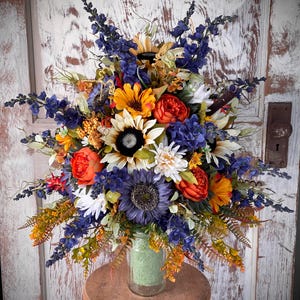 May include: A vibrant floral arrangement featuring sunflowers, orange peonies, and blue delphiniums. The bouquet is arranged in a clear glass jar filled with green material, placed on a small wooden stool. The background is a weathered white door.