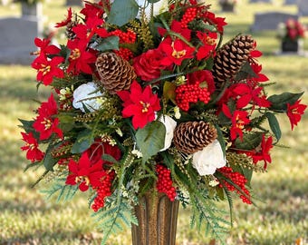 Könnte beinhalten: Ein Blumenarrangement mit roten Weihnachtssternen, weißen Rosen, braunen Tannenzapfen und roten Beeren in einer hohen, geriffelten, bronzefarbenen Vase. Das Arrangement enthält grünes Blattwerk und steht auf einem Granitsockel.