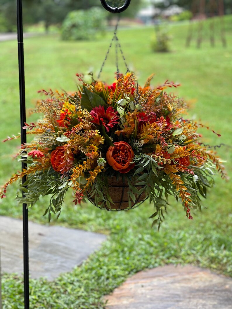 K&ouml;nnte beinhalten: Ein H&auml;ngekorb-Arrangement mit einer dichten Ansammlung k&uuml;nstlicher Blumen und Bl&auml;tter in den Farben Dunkelorange, Rost, Gelb und Gr&uuml;n. Das Gesteck besteht aus runden Bl&uuml;ten, schlanken Blattstielen und strukturierten Gr&auml;sern in einem Drahtkorb.