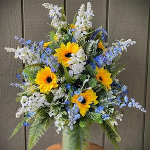 May include: A bouquet of yellow sunflowers, blue delphiniums, and white flowers in a clear glass vase. The vase is sitting on a wooden stool.