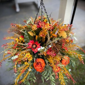 K&ouml;nnte beinhalten: Ein h&auml;ngendes Blumenarrangement mit einer dichten Ansammlung k&uuml;nstlicher Blumen und Bl&auml;tter in Herbstfarben. Das Gesteck enth&auml;lt leuchtend orangefarbene Pfingstrosen, rote Sonnenblumen, gelbe G&auml;nsebl&uuml;mchen sowie verschiedene gr&uuml;ne und rostfarbene Bl&auml;tter.