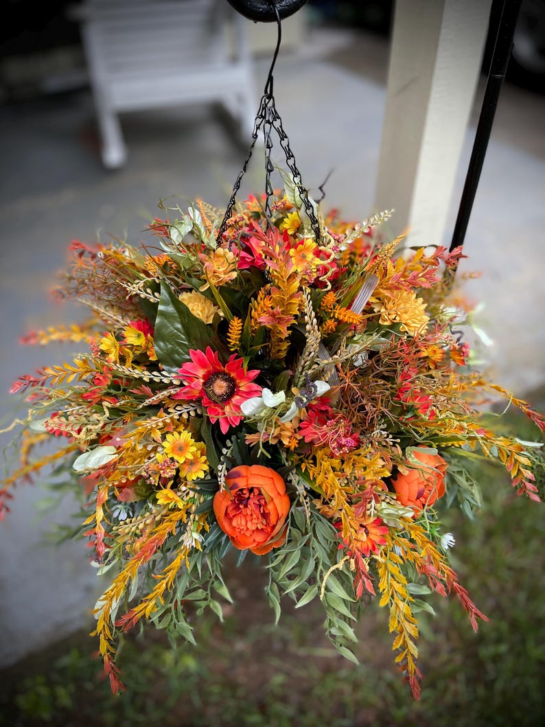 K&ouml;nnte beinhalten: Ein h&auml;ngendes Blumenarrangement mit einer dichten Ansammlung k&uuml;nstlicher Blumen und Bl&auml;tter in Herbstfarben. Das Gesteck enth&auml;lt leuchtend orangefarbene Pfingstrosen, rote Sonnenblumen, gelbe G&auml;nsebl&uuml;mchen sowie verschiedene gr&uuml;ne und rostfarbene Bl&auml;tter.