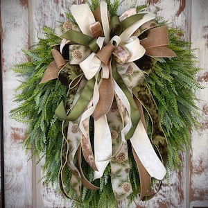 May include: A green, brown, and white wreath with a large bow made of burlap, velvet, and satin ribbon. The wreath is decorated with pine cones and is perfect for a rustic or farmhouse style decor.