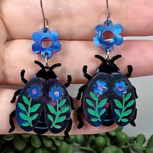 May include: A pair of dangle earrings featuring a black beetle design with blue and green floral accents. Each earring has a blue flower at the top and is suspended from a silver-colored hook. The earrings are displayed against a neutral background.