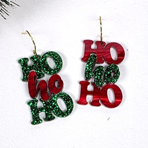 May include: A pair of dangle earrings featuring the text HO HO HO in alternating red and green colors. The letters are crafted from a mix of solid red and glittery green material, suspended from gold-toned metal earring hooks.