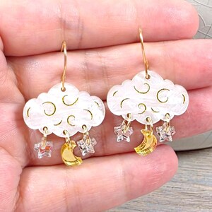May include: Cloud-shaped earrings with gold-colored hooks. The white clouds have curved lines and are adorned with gold crescent moons, clear star-shaped charms, and small square charms. The earrings are held in a hand.