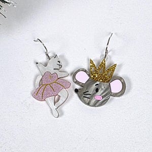 May include: Two whimsical earrings: one a white cat in a pink tutu, the other a gray mouse with a gold crown. The earrings are set against a white background with festive greenery, suggesting a holiday theme. The cat earring is approximately 1.5 inches tall.