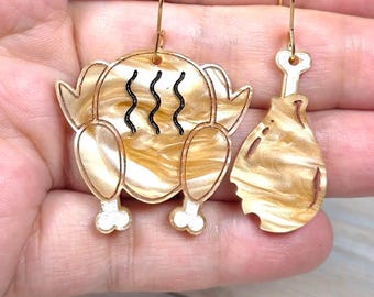May include: A pair of mismatched dangle earrings featuring a roasted turkey and a single turkey leg. Both pieces are crafted from a tan and cream marbled acrylic material with gold-toned metal hooks and black wavy steam lines on the turkey.