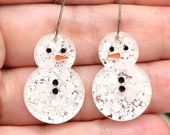 May include: A pair of snowman-shaped dangle earrings featuring a white glittery finish, black dot eyes and buttons, and a small orange carrot-shaped nose. The earrings are attached to silver-toned metal hooks for pierced ears.