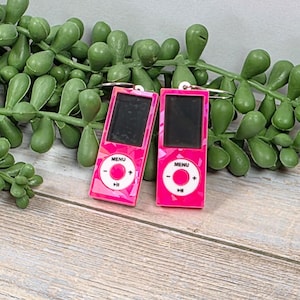 May include: Two pink iPod-shaped earrings with a geometric pattern. Each earring has a black screen, a white center button, and the word "MENU" above the button. The earrings are displayed on a wooden surface with green plant stems.