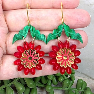 May include: A pair of red flower earrings with gold accents and green leaf details. The earrings have gold-colored hooks. The flower petals are a vibrant red, and the center of the flower has a gold design.