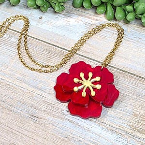 May include: A gold-tone chain necklace with a large, layered red flower pendant. The flower has a gold-colored center and is set against a light wood-grain background. Green foliage is visible in the background.