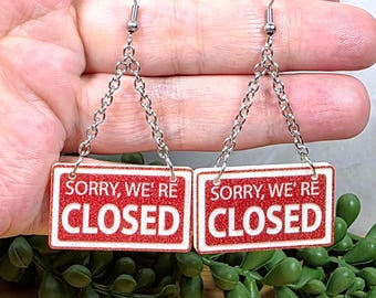 May include: A pair of earrings featuring small, rectangular signs with the words "SORRY, WE'RE CLOSED" in red and white. The earrings hang from silver-colored chains and hooks. The background is a hand and greenery.