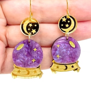 May include: A pair of dangle earrings featuring a purple marbled resin crystal ball charm with gold star accents. Above the crystal ball is a circular black charm with a gold crescent moon and stars, suspended from gold-toned metal hooks.