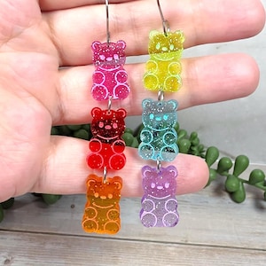 May include: Rainbow-colored gummy bear earrings. Each earring features a vertical row of three gummy bear charms in pink, red, orange, yellow, blue, and purple. The charms have a glittery finish and are suspended from silver-tone ear wires.