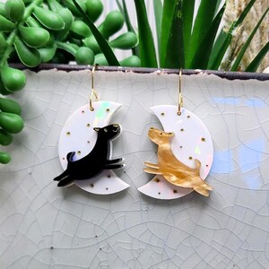 May include: A pair of dangle earrings featuring white crescent moon charms with iridescent finishes and small gold star accents. One earring displays a black dog silhouette, while the other features a tan marbled dog silhouette, both suspended from gold-toned hooks.