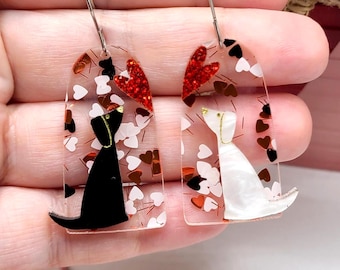 May include: A pair of arch-shaped dangle earrings featuring a clear acrylic base with scattered white and red heart shapes. One earring displays a solid black dog silhouette, while the other features a white pearlescent dog silhouette with silver hooks.
