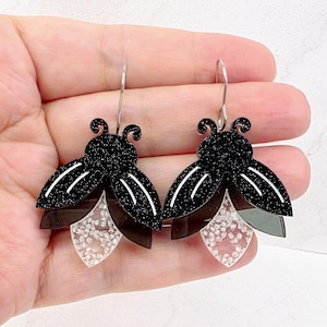 May include: A pair of dangle earrings shaped like fireflies, featuring black glitter acrylic bodies with white line details and translucent, textured clear acrylic lower sections. The earrings are attached to silver-toned metal ear wires.