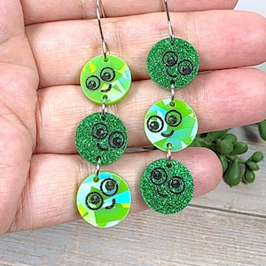 May include: Green and glittery dangle earrings with a whimsical design. Each earring features three circular charms with smiling faces. The top and bottom charms have a geometric pattern, while the middle charm has a glittery texture.