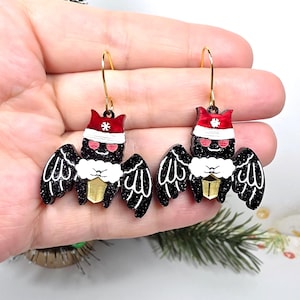 May include: A pair of dangle earrings featuring black glittery bats wearing red and white Santa hats with a white snowflake detail. The bats have pink eyes, white fur collars, and hold a small gold-colored lantern. Gold-toned metal hooks.