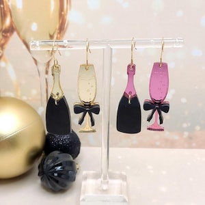 May include: Champagne-themed earrings in black, gold, and pink. The earrings are shaped like champagne bottles and glasses, with black bows. The earrings are displayed on a clear stand, with a gold ornament and champagne glasses in the background.