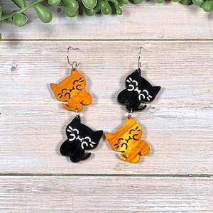 May include: A pair of cat-shaped dangle earrings. One earring features a black cat above an orange cat, and the other features an orange cat above a black cat. Each cat has a smiling face and small white details.