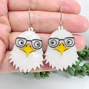May include: A pair of dangle earrings featuring white eagle heads with yellow beaks and black-rimmed glasses. The eagle faces are crafted with a pearlescent finish and are suspended from silver-toned metal hooks for pierced ears.