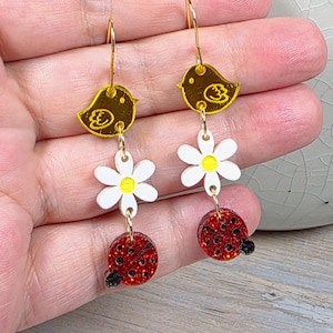 May include: A pair of dangle earrings featuring a gold bird, white daisy, and red ladybug design. The earrings have gold-colored hooks and are made of acrylic. The ladybug has black spots and a black head.
