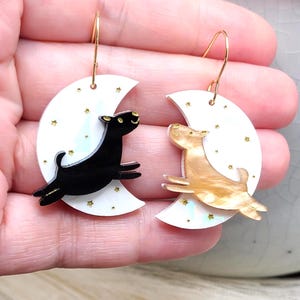 May include: A pair of dangle earrings featuring white crescent moon shapes decorated with small gold stars. One earring displays a black dog silhouette, while the other features a tan, marbled dog silhouette. Both have gold-toned metal hooks.