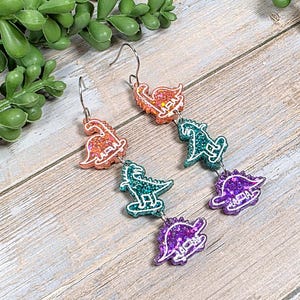 May include: Dinosaur-themed dangle earrings. Each earring features three glittery dinosaur shapes in orange, teal, and purple, connected by silver-colored metal. The earrings have a hook closure and are set against a wooden background.